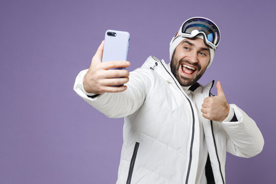 Cheerful skier man in white jacket ski goggles mask doing selfie shot on mobile phone showing thumb up spend weekend winter in mountains isolated on purple background. People lifestyle hobby concept.