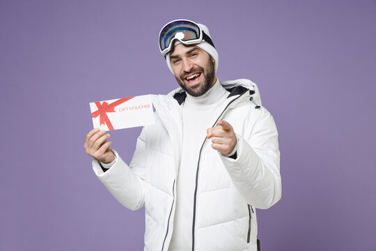 Funny skier man in white jacket ski goggles mask hold gift certificate pointing index finger on camera spend weekend winter in mountains isolated on purple background. People lifestyle hobby concept.