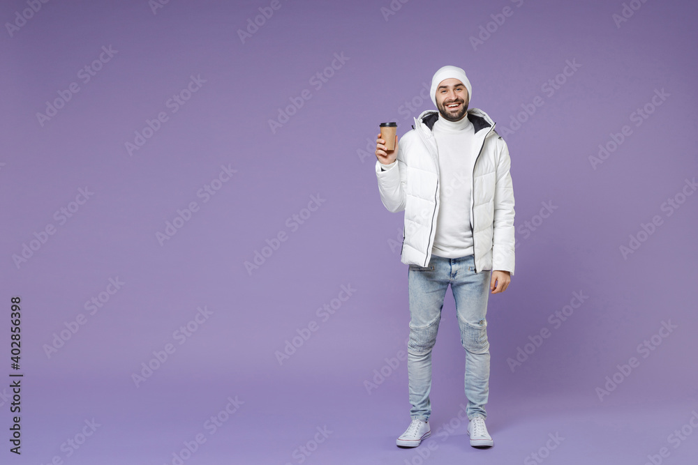 Wall mural Full length of smiling attractive man in warm white windbreaker jacket hat hold paper cup of coffee or tea isolated on purple background studio portrait. People lifestyle cold winter season concept.