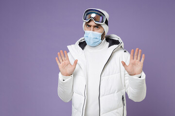 Worried skier man in warm white windbreaker jacket ski goggles face mask safe from coronavirus virus covid-19 rising hands up weekend in mountains isolated on purple background. People hobby concept.