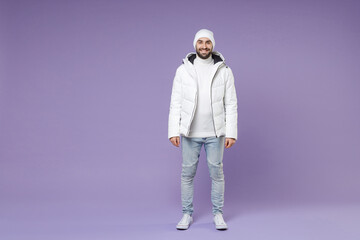 Full length of smiling attractive man in warm white padded windbreaker jacket hat standing looking camera isolated on purple background studio portrait. People lifestyle cold winter season concept. © ViDi Studio