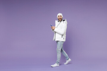 Full length side view surprised man in warm white windbreaker jacket hat using mobile cell phone typing sms message isolated on purple background studio portrait. People lifestyle cold season concept.