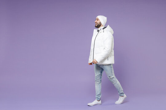 Full length side view of cheerful man in warm white windbreaker jacket hat walking going looking aside isolated on purple color background studio portrait. People lifestyle cold winter season concept.