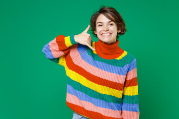 Smiling young brunette woman 20s years old wearing casual colorful sweater standing doing phone gesture like says call me back looking camera isolated on bright green color background studio portrait.
