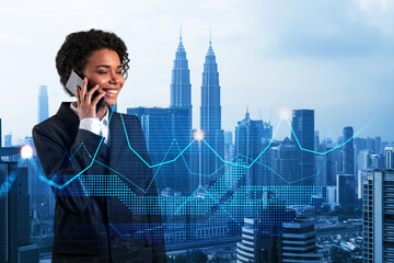Attractive black businesswoman pensively processing conference call by phone to find financial solution for venture capital project. Hologram chart over Kuala Lumpur city background