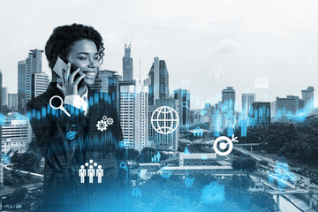 Attractive black businesswoman trying to find new ideas using smartphone as a modern tool to solve problems at research and development department. Fintech hologram icons over Kuala Lumpur.