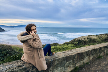 woman talking on the smartphone sitting looking at the views of the sea and the landscape in winter.