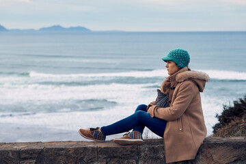 woman with hat and coat looking at the seascape and cliffs in winter on a cloudy day carrying her newborn baby. concept go out in winter