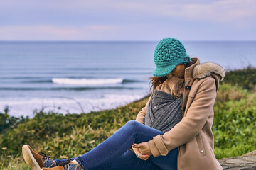 portrait of woman with hat and coat looking at the seascape and cliffs in winter on a cloudy day carrying her newborn baby. concept go out in winter