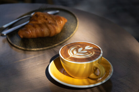 Cup of coffee with latte art with croissants on wooden table and morning sunlight with shadow through from window. Beautiful meal with warm sunlight.