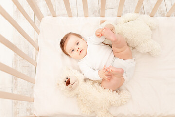 the baby is six months old in a crib in a white bodysuit with a Teddy bear