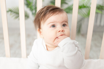 happy baby sitting in the crib in a white bodysuit and sucking his finger