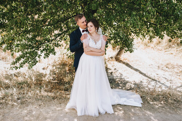 Stylish groom in a blue suit and a beautiful curly-haired bride in a white dress are hugging on the background of nature, in a green park. Wedding portrait of newlyweds in love.