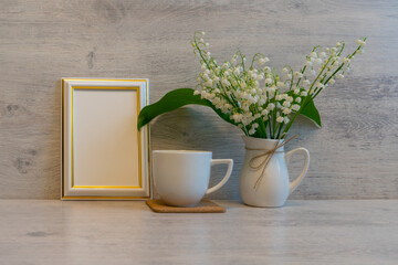 Bouquet of fresh lily of the valley flowers with tea or coffee cup and empty photoframe on light wooden background. Beautiful spring still life, mock up for greeting card, good morning concept