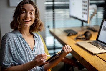 Young woman dressed casually having some creative work, drawing on a digital tablet, sitting at the cozy and stylish home office