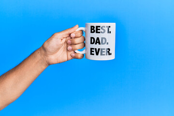 Hand of hispanic man holding best dad ever coffee cup over isolated blue background.