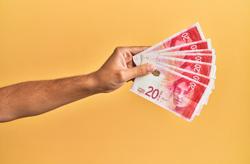Hand of hispanic man holding israel 20 shekels banknotes over isolated yellow background.