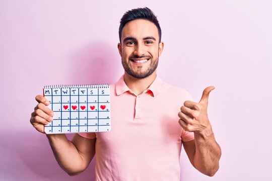 Handsome romantic man with beard holding calendar with red hearts over pink background smiling happy and positive, thumb up doing excellent and approval sign