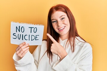 Young red head girl holding no to suicide paper smiling happy pointing with hand and finger