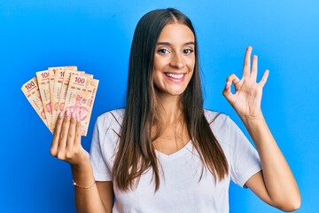 Young hispanic woman holding mexican pesos doing ok sign with fingers, smiling friendly gesturing...