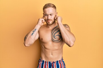 Fototapeta premium Young caucasian man wearing swimwear shirtless covering ears with fingers with annoyed expression for the noise of loud music. deaf concept.