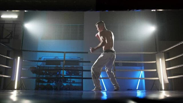 Male taekwondo fighter training at dark smoky ring with spotlights. Shadow fight. Kickboxer training alone. Shirtless fighter. Full length in 4K, UHD
