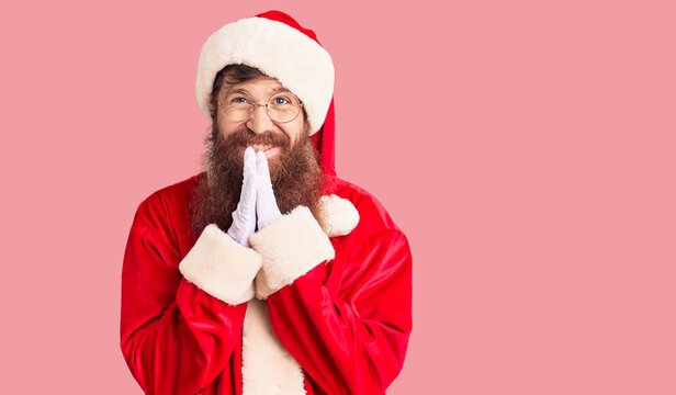 Handsome young red head man with long beard wearing santa claus costume praying with hands together asking for forgiveness smiling confident.