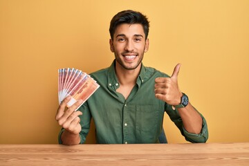 Young handsome man holding norwegian krone banknotes smiling happy and positive, thumb up doing...