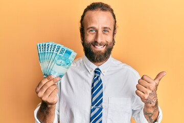 Handsome business man with beard and long hair holding 100 brazilian real banknotes pointing thumb up to the side smiling happy with open mouth