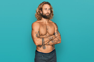 Handsome man with beard and long hair standing shirtless showing tattoos looking to the side with arms crossed convinced and confident
