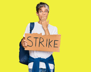 Young hispanic man holding strike banner cardboard covering mouth with hand, shocked and afraid for...