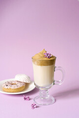 Beautiful dalgona drink a frothy coffee in a transparent mug and flowers of lilac on a purple background. Sweets on a plate: Donat and tender marshmallows. Copy space