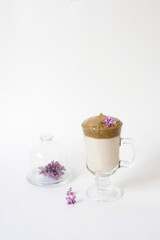 Dalgona coffee in a transparent mug with a lilac flower on a white background and a sprig of lilac lying on a serving plate