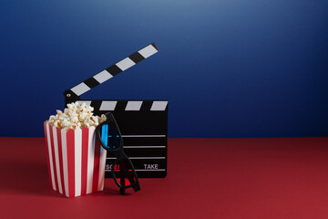 Cinema clapperboard, colourful 3d glasses and red striped bucket with popcorn on red and blue...