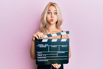 Beautiful caucasian blonde girl holding video film clapboard puffing cheeks with funny face. mouth...