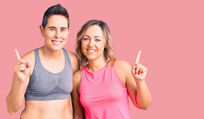 Couple of women wearing sportswear with a big smile on face, pointing with hand finger to the side looking at the camera.