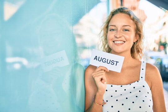 Young beautiful caucasian woman with blond hair smiling happy outdoors on a summer day holding August word