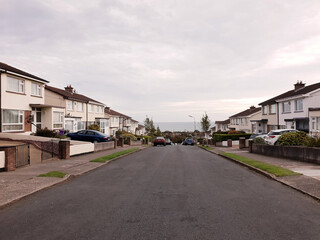 Fototapeta na wymiar Suburban street with modern Irish houses and parked cars in seaside town. The Irish Sea visible on the horizon. Quiet road in residential area.