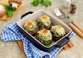 Baked brown mushrooms stuffed with spinach and cheese, with breadcrumbs and cheese crust in a black portioned pan for baking on a gray concrete background. Champignon recipes.
