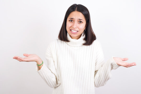 Clueless Young brunette woman wearing white knitted sweater against white background shrugs shoulders with hesitation, faces doubtful situation, spreads palms, Hard decision