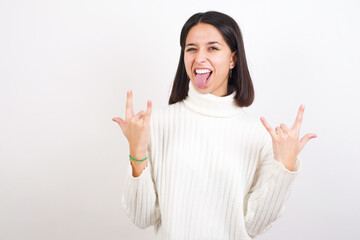 Young brunette woman wearing white knitted sweater against white background making rock hand...
