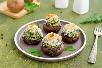 Baked brown mushrooms stuffed with spinach and cheese, with breadcrumbs and cheese crust on a brown plate on a green concrete background. Champignon recipes.