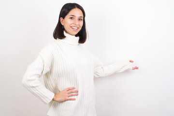 Young brunette woman wearing white knitted sweater against white background feeling happy and cheerful, smiling and welcoming you, inviting you in with a friendly gesture