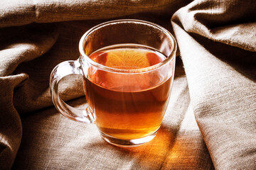 Black tea in transparent glass. Golden color tea. Brown hot beverage. Tea mug on linen fabric. Morning hot drink on vintage canvas.