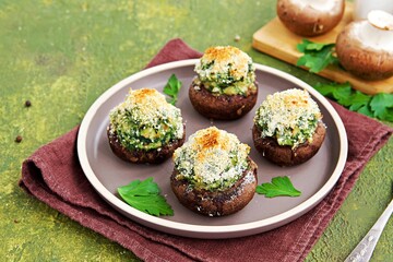 Baked brown mushrooms stuffed with spinach and cheese, with breadcrumbs and cheese crust on a brown plate  on a green concrete background. Champignon recipes.