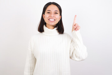 Young brunette woman wearing white knitted sweater against white background showing and pointing up with finger number one while smiling confident and happy.