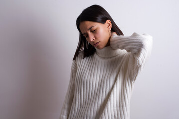 Young brunette woman wearing white knitted sweater against white background Suffering of neck ache injury, touching neck with hand, muscular pain