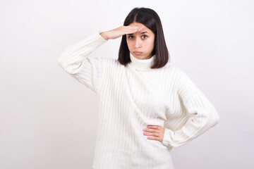 Young brunette woman wearing white knitted sweater against white background having problems, worried and stressed holds hand on forehead.