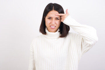 Young brunette woman wearing white knitted sweater against white background pointing unhappy at pimple on forehead, blackhead  infection. Skincare concept.