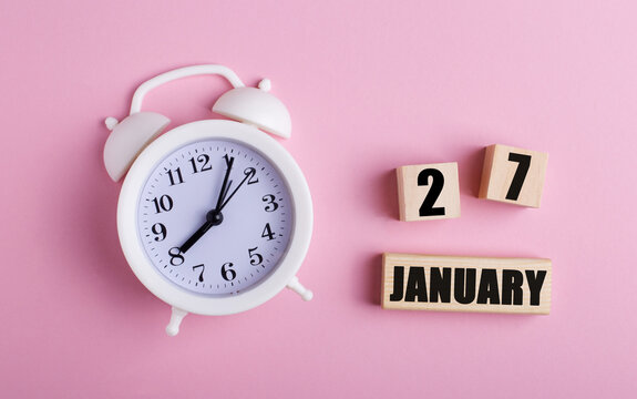 On a pink background, a white alarm clock and wooden cubes with the date of JANUARY 27